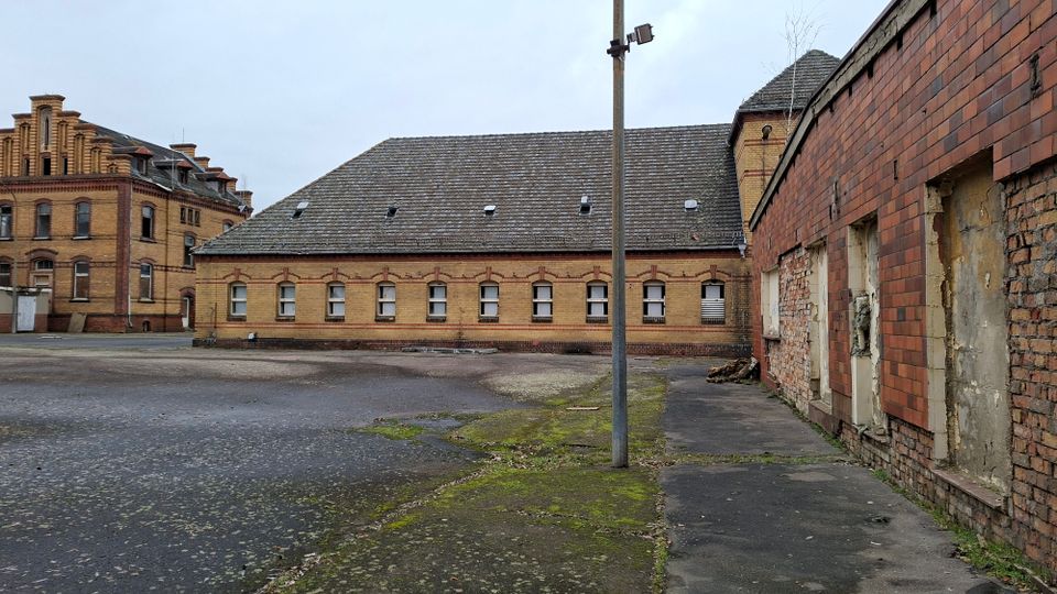 Innenhof eines alten Backstein-Industriekomplexes mit langem, niedrigem Gebäude und Satteldach sowie einem mehrstöckigen Ziegelbau im Hintergrund. Im Vordergrund liegt eine große asphaltierte Fläche mit Moosstellen und einem hohen Mast; rechts ist eine teilweise beschädigte Ziegelwand mit zugemauerten Öffnungen zu sehen.
