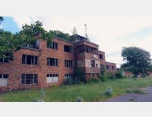 Backsteingebäude in verfallenem Zustand, mit zerbrochenen Fenstern und überwucherten Pflanzen an der Fassade, umgeben von Gras und Bäumen; im Hintergrund ein Himmel mit vereinzelten Wolken.