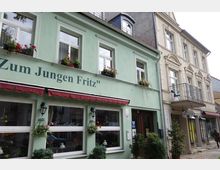 Straßenansicht eines zweistöckigen grünen Gebäudes mit der Aufschrift „Zum Jungen Fritz“ und Blumenkästen an den Fenstern, daneben ein beiges Gebäude mit Balkon und dekorativer Fassade. Im Vordergrund Gehweg mit kleineren Pflanzen.