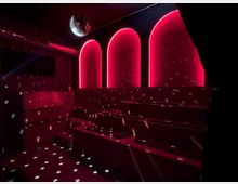 Ein dunkler Raum mit mehreren gestuften Sitzreihen und roten Neonlichtern, die drei bogige Nischen im Hintergrund umrahmen. Eine Discokugel hängt an der Decke und wirft Lichtpunkte auf die Wände und Stufen.