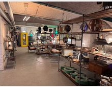 Industrie-ähnlicher Innenraum mit gewölbter Backstein-Decke, Regalen voller Vintage-Gegenstände wie Lampen, Elektrogeräte und Schilder; im Hintergrund eine gelbe Telefonzelle und dekorative Beleuchtung. Die Wände bestehen aus weiß gestrichenem Mauerwerk, und der Boden ist aus Beton.