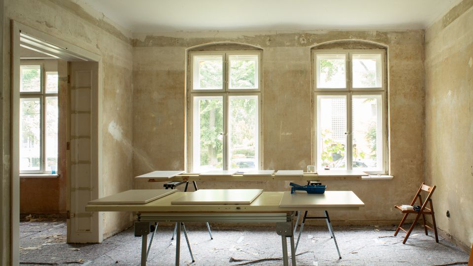 Unrenovierter Innenraum mit freigelegten, rauen Wänden und zwei großen, weiß gerahmten Sprossenfenstern, durch die Tageslicht einfällt. Im Raum stehen mehrere Arbeitstische mit Platten und Werkzeug, daneben ein Durchgang zu einem weiteren Zimmer und ein einzelner Stuhl am Rand.