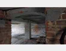 Ein leerer Kellerraum mit rohen Ziegelwänden, Betonbögen und Boden aus Ziegelsteinen. In der Mitte des Raumes stehen gemauerte Säulen, und Holzpaletten liegen am Boden.