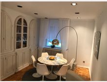 Esszimmer in heller, moderner Einrichtung mit einem runden Marmortisch, weißen Stühlen und einer geschwungenen Bogenlampe. An der Wand befindet sich eine weiße Einbauschrankwand, daneben ein Fenster mit weißen Vorhängen.