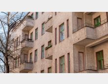 Wohnhausfassade mit beige verputzten Wänden, zahlreichen grünen Fenstern und Balkonen mit einfachen Metallgeländern; im Vordergrund ein kahler Baum ohne Laub.