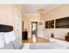 Schlafzimmer in einem Altbau mit hohen Decken und Parkettboden. Es sind ein Bett mit grauem Kopfteil, eine schwarze Kommode, zwei großformatige Wandbilder und links eine geöffnete Tür sichtbar, die in einen weiteren Raum mit Fenster führt.