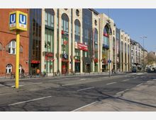 Straßenszene mit moderner Einkaufszeile, bestehend aus einem Gebäude mit großen Glasfronten und dekorativen Säulen; im Vordergrund ein gelbes U-Bahn-Schild der Linie U9 und Fahrradständer entlang des Bürgersteigs. Auf der linken Seite ist ein rotes Backsteingebäude zu sehen, rechts weitere Wohn- und Geschäftsgebäude.