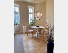 Essbereich in einem hellen Raum mit Holzboden, einem runden weißen Esstisch, vier Holzstühlen mit hellen Sitzkissen und einer farbenfroh gemusterten Hängelampe. An den Fenstern stehen Zimmerpflanzen, rechts im Vordergrund ein großer blauer Vase mit Zweigen.