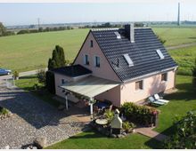 Einfamilienhaus mit rosa Fassade und schwarzem Satteldach, umgeben von einem großen Garten mit Blumenbeeten und Rasen. Im Vordergrund ist eine gepflasterte Einfahrt und eine überdachte Terrasse zu sehen; im Hintergrund erstrecken sich weite Felder mit Windrädern.