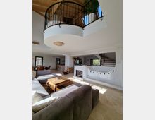 Moderne Wohnzimmereinrichtung mit offener Gestaltung. Sichtbar sind eine graue Sofalandschaft, ein Holztisch, ein Kamin in einer weißen Wand mit Rundungselementen, Holzstapel, sowie eine freitragende Treppe aus Holz; darüber ein offener Balkon mit schwarzem Geländer und dekorativer Zimmerpflanze.