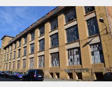 Fabrikgebäude aus gelbem Backstein mit großen, teilweise beschädigten Fenstern und sichtbaren Graffiti an der Wand; vor dem Gebäude parken mehrere Autos. Der Himmel ist blau und klar.