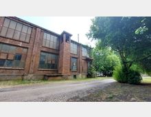 Backstein-Gebäude einer alten Fabrik mit großen, teilweise beschädigten Fenstern und einem Außengerüst, daneben ein Pflasterweg und dichte Bäume im Hintergrund.