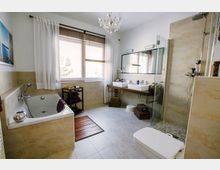 Modernes Badezimmer mit hellen Fliesenwänden, einer eingebauten Badewanne links, einem Waschtisch mit zwei Waschbecken und großem Spiegel in der Mitte sowie einer bodengleichen Dusche mit Glasabtrennung rechts. Ein Fenster mit weißen Vorhängen und Jalousien sorgt für Tageslicht.