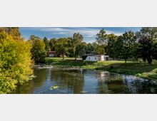 Ein Fluss mit klarem Wasser, umgeben von grünen Bäumen und einer Wiese, auf der ein weißes und ein braunes Gebäude stehen. Im Hintergrund sind weitere Bäume und ein blauer Himmel mit leichten Wolken zu sehen.