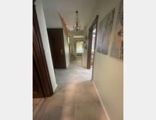 Flur eines Wohnhauses mit einem hellen Bodenbelag, links eine dunkle Holztür und rechts an der Wand hängende Bilder. Im Hintergrund führen eine Treppe nach oben sowie eine Türöffnung ins Freie mit Blick auf grüne Pflanzen. Eine Hängeleuchte ist an der Decke angebracht.