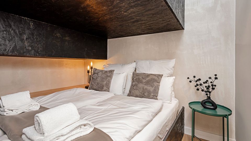 Modernes Schlafzimmer mit Doppelbett und weißer Bettwäsche unter einer markanten, dunklen, kantigen Deckenverkleidung. Neben dem Bett steht ein kleiner grüner Beistelltisch mit Vase, dazu eine schlichte Wandlampe und helle, glatte Wände.