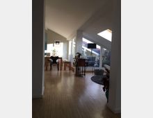 Ein modernes Wohnzimmer mit schräger Decke und großen Dachfenstern, die viel Tageslicht einlassen. Der Raum ist mit Holzboden ausgelegt und enthält eine Sitzgruppe, einen kleinen Tisch mit Accessoires und eine Essbereich-Ecke mit Holz- und Glasdetails.