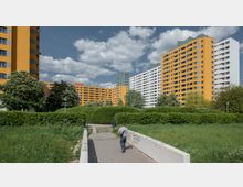 Mehrstöckige Wohngebäude in gelben und weißen Farben, umgeben von grünen Bäumen und Hecken in einer parkähnlichen Umgebung. Im Vordergrund führt ein gepflasterter Weg mit einer niedrigen Brüstung durch eine Wiese.