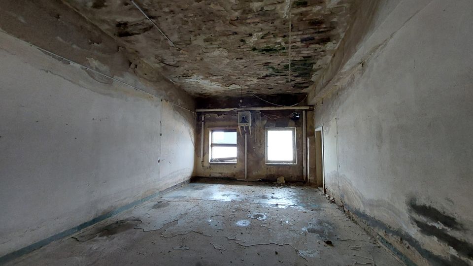 Ein leerer, heruntergekommener Raum mit abblätterndem Putz an den Wänden sowie Decken- und Wandbeschädigungen. Zwei Fenster an der Stirnwand lassen Tageslicht in den Raum, während der Boden Spuren von Feuchtigkeit und Abnutzung zeigt.