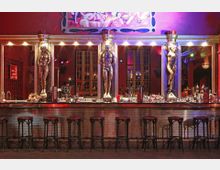 Eine stilvolle Bar mit einer langen Theke, dekoriert mit goldenen Skulpturen menschlicher Figuren zwischen Spiegeln. Die Wand dahinter ist in Rot gehalten, leuchtende Akzente und eine künstlerische Illustration sind sichtbar.