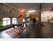 Ein moderner Besprechungsraum in einem industriellen Stil mit Betonwänden, großen bogenförmigen Fenstern und einem dunklen Boden. Der Raum ist mit Holztischen, Stühlen, bunten Teppichen und zahlreichen Pflanzen dekoriert; eine Holzwand und eine Präsentationsfläche befinden sich im Hintergrund.