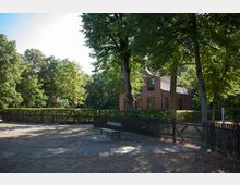 Roter Backsteinbau mit Satteldach, umgeben von hohen Bäumen, einem Holzzaun und einer Hecke, im Vordergrund ein gepflasterter Platz mit einer Parkbank.