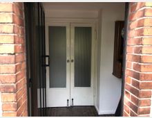 Eingangsbereich eines Gebäudes mit roten Backsteinwänden, einer weißen Doppeltür mit geriffeltem Glas und einem braunen Fliesenboden. Rechts an der Wand ist eine hölzerne Wanduhr angebracht.