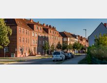 Reihenhauszeile mit roten Backsteinfassaden und Satteldächern, entlang einer Straße mit parkenden Autos und vereinzelten Bäumen, in einer ruhigen städtischen Wohngegend bei Sonnenschein.