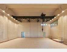 Großer, heller Innenraum mit hohen Decken, cremefarbenen Wänden und einem schwarz lackierten Trägergerüst mit einem Kettenzug. Der Boden besteht aus einem karoförmigen Fliesenmuster, und seitlich befinden sich kleine Fenster sowie Türen, die in andere Räume führen.