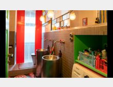 Kleines, modernes Badezimmer mit industriellem Design, gekennzeichnet durch zwei runde Edelstahllavabos und grau geflieste Wände. Im Hintergrund befinden sich große Fenster mit roten halbtransparenten Vorhängen, während an der Seite ein Regal mit bunten Kisten und Pflegeprodukten zu sehen ist.