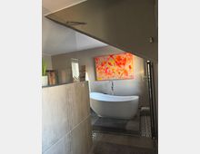 Modernes Badezimmer mit freistehender, weißer Badewanne auf erhöhtem Podest. An der Wand darüber hängt ein farbenfrohes, abstraktes Gemälde in Orange- und Pinktönen, umgeben von Fliesen mit geometrischen Mustern und schlichten grauen Wand- und Bodenfliesen.