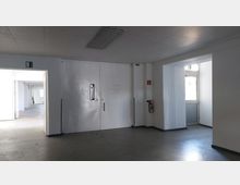 Ein industrieller Innenraum mit weiß gestrichenen Wänden, einer großen, weißen Doppeltür mit kleinem Sichtfenster und einer Notausgangstür mit Oberlicht. An der Wand hängt ein Feuerlöscher, und durch geöffnete Türen sind angrenzende Räume zu sehen.