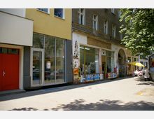Straßenszene mit mehrstöckigen Gebäuden, darunter eine gelbe Fassade mit großen Glasfenstern und eine ältere, graue Hausfassade mit Schaufenstern, die mit Plakaten beklebt sind. Rechts ein Bürgersteig mit Baum und einem kleinen Cafébereich mit gelbem Sonnenschirm und Tischen.