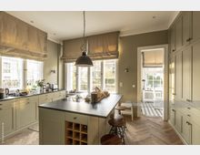 Helle, moderne Küche mit hellgrünen Schränken, einer schwarzen Arbeitsplatte und einer großen Kücheninsel mit Barhockern. Große, sprossengeteilte Fenster lassen viel Tageslicht herein, während im Hintergrund ein angrenzender Essbereich mit schwarz-weißem Fliesenboden sichtbar ist.