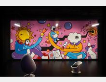 Ein buntes Wandgemälde in einem modernen Innenraum zeigt surrealistische Figuren vor einem rosa Hintergrund mit Sternen und Planeten; im Vordergrund stehen zwei futuristisch geformte Stühle.