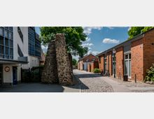Straßenszene mit gepflastertem Gehweg, links ein modernes Gebäude mit weißen Wänden und blauen Fensterelementen, rechts Backsteingebäude, darunter historische Ruinen aus Natursteinen, sowie ein Baum im Hintergrund. Der Himmel ist teilweise bewölkt, Fahrräder stehen an der Hauswand.