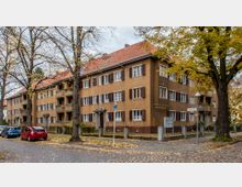 Mehrstöckiges Wohnhaus aus der Vorkriegszeit mit brauner Fassade, weißen Fensterrahmen und dunkelbraunen Fensterläden. Vor dem Gebäude stehen große Bäume mit Herbstlaub, während Autos am gepflasterten Straßenrand parken.