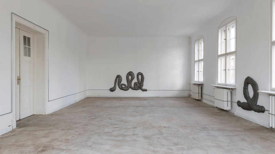 Ein leerer, heller Raum mit weißen Wänden und großen, rechteckigen Fenstern, durch die Tageslicht einfällt. Der Boden ist aus Beton, und an der Wand sowie unter den Fenstern sind Heizkörper angebracht. An zwei Wänden befinden sich skulpturale, dunkle Röhrenobjekte.