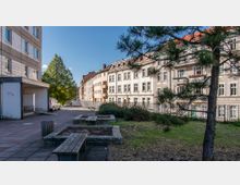 Innenhof mit gepflasterten Wegen, Sitzbänken und kleinen bepflanzten Beeten; im Hintergrund sind Wohnhäuser mit hellen Fassaden und Balkonen sowie ein Baum sichtbar, rechts ein Nadelbaum im Vordergrund.