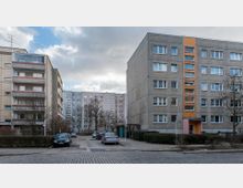 Straßenansicht mit Wohnblöcken im Plattenbaustil, umgeben von kahlen Bäumen und einem gepflasterten Gehweg. Im Vordergrund sind parkende Autos auf einem kleinen Parkplatz zu sehen, während der Himmel wolkig ist.