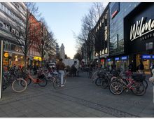 Städtische Einkaufsstraße mit modernen Geschäften und Passanten in einer Fußgängerzone. Zahlreiche Fahrräder sind auf einem befestigten Platz abgestellt, umgeben von Gebäuden mit Schaufenstern, darunter ein Einkaufszentrum mit der Aufschrift „Wilmers“.