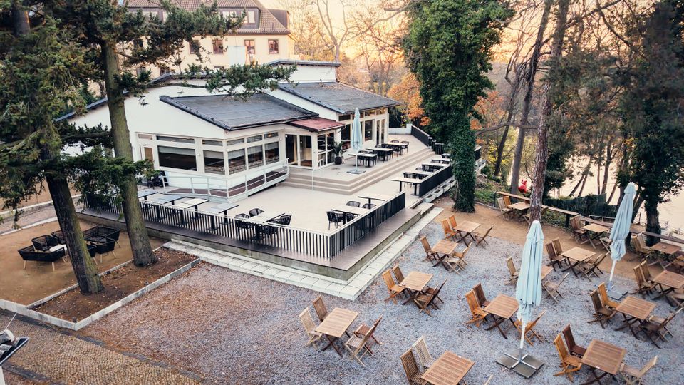 Outdoor dining area surrounded by tall trees, featuring wooden tables and chairs arranged on a gravel surface, alongside a modern white building with large windows and a raised terrace. The setting is within a wooded landscape, with warm light suggesting a sunset in the background.