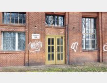 Backsteinfassade eines alten Gebäudes mit der Bezeichnung „Gebäude 11“ auf einem Schild. Zu sehen sind ein gelbes Holztor, mehrere hohe Fenster mit Sprossen sowie Graffiti an den Wänden.