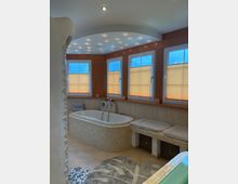 Modernes Badezimmer mit breiter Badewanne, gefliesten Wänden und einer Sitzbank mit Kissen und Körben darunter. Drei große Fenster mit Plissees lassen Tageslicht herein, während eine Decke mit integrierten Spotlichtern für zusätzliche Beleuchtung sorgt.