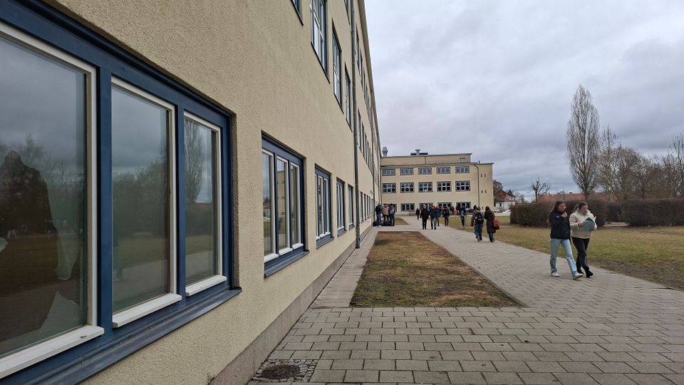 Langer, mehrstöckiger Gebäudekomplex mit hell verputzter Fassade und blau gerahmten Fenstern entlang eines gepflasterten Gehwegs. Rechts daneben liegen Rasenflächen mit kahlen Bäumen; im Hintergrund steht ein weiterer flacher Gebäudetrakt unter bedecktem Himmel.