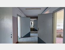 Ein leerer, verlassener Flur mit mehreren geöffneten grauen Türen, die in andere Räume führen. Die Wände sind teilweise gestrichen, der Boden besteht aus grauen Fliesen, und rechts ist ein Bereich mit gefliesten Wänden und einer Arbeitsplatte sichtbar.