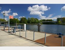 Steg an einem Flussufer mit einem gelben Pflasterboden, einem Geländer und mehreren Pollern; im Hintergrund eine Brücke, Bäume und ein Gebäude, bei klarem Himmel mit wenigen Wolken.