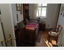 Kleines Schlafzimmer mit Holzboden, einem Einzelbett mit buntem Patchwork-Bettüberwurf, einer Holzbücherwand, mehreren gemusterten Kissen und einem Nachttisch mit Tischlampe. Durch ein großes Fenster fällt Tageslicht herein, und es befindet sich ein schlichter Holzstuhl im Raum.