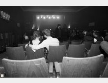 Ein Zuschauerraum mit Holzstühlen in einem dunklen Saal, im Vordergrund ein Paar, das sich ansieht, während es in den hinteren Reihen weitere Personen und eine Bühne mit eingeschalteten Scheinwerfern gibt.