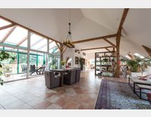 Geräumiges Loft mit offenen Holzbalken, großen Fenstern und einem Terrassenbereich mit Glasüberdachung. Der Raum ist mit Fliesenboden, einem Essbereich, Pflanzen und Regalen eingerichtet, die eine luftige und helle Atmosphäre schaffen.